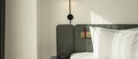 Una cama bien hecha con ropa blanca, una mesita de noche pequeña con una lámpara y un reloj de pared.
