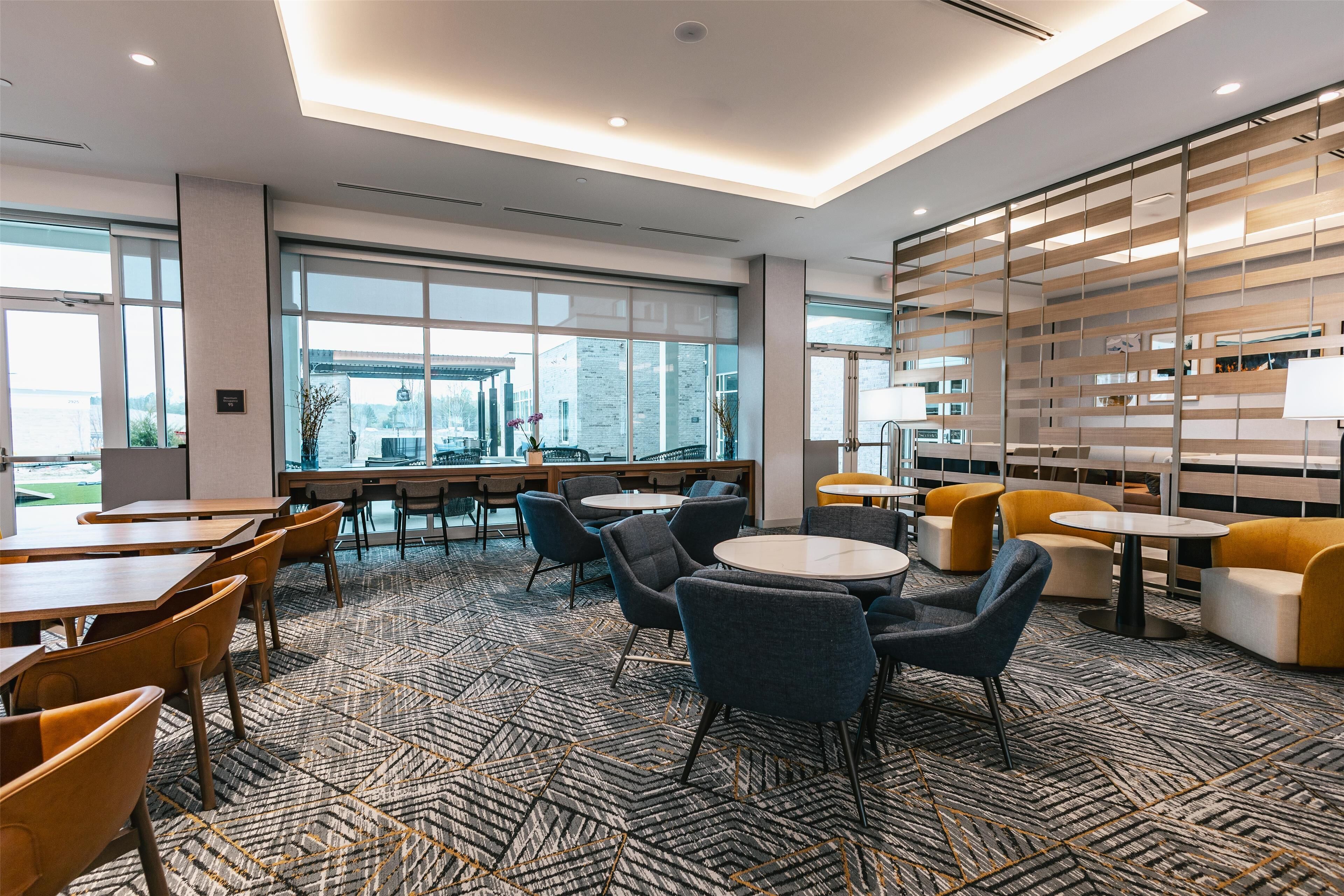 A modern indoor space with tables and chairs, large windows, and a patterned carpet.