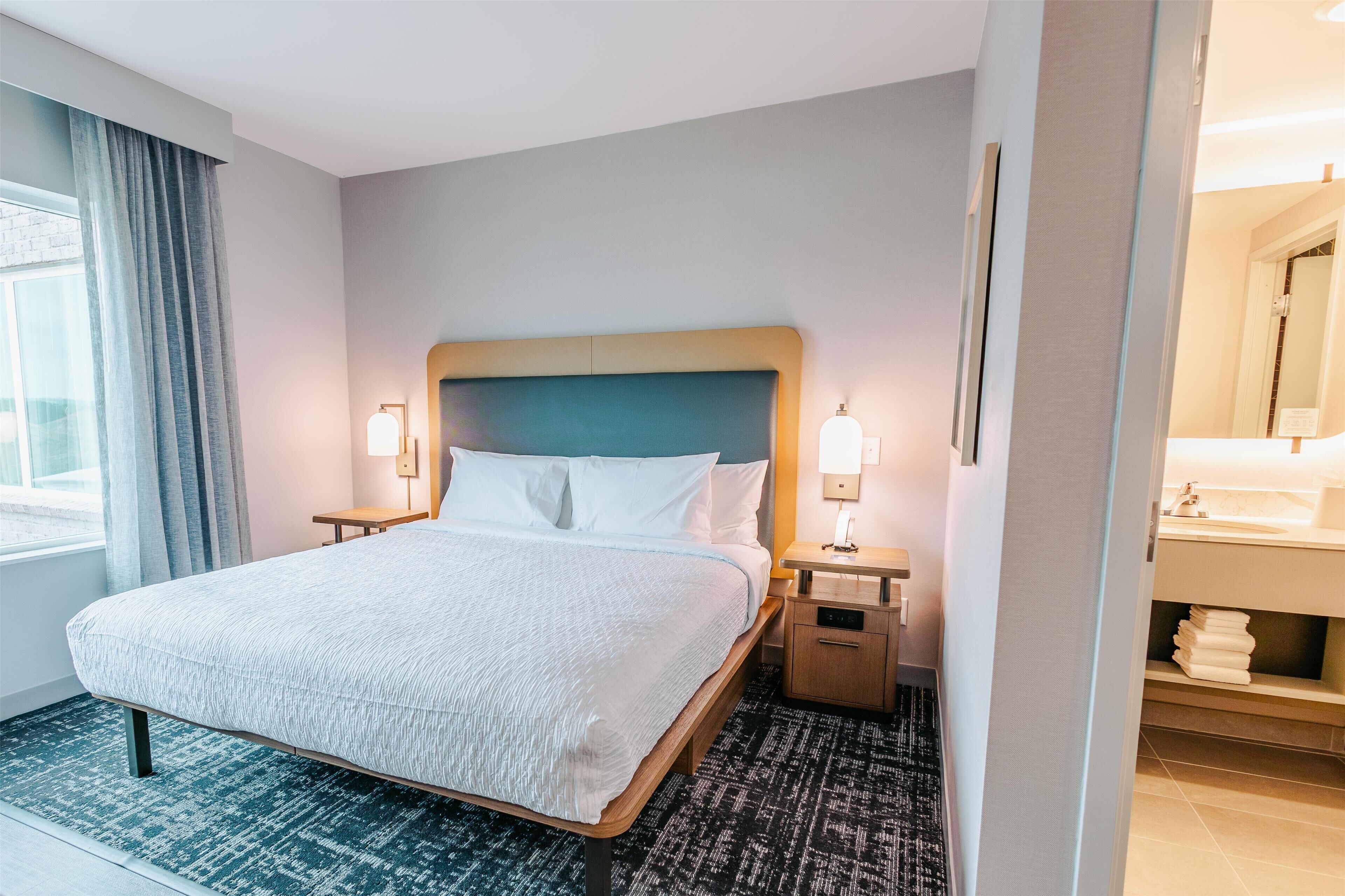 Una habitación de hotel con una cama grande, dos mesitas de noche con lámparas, una mesita auxiliar, un baño con lavabo y espejo, y una ventana con cortinas.