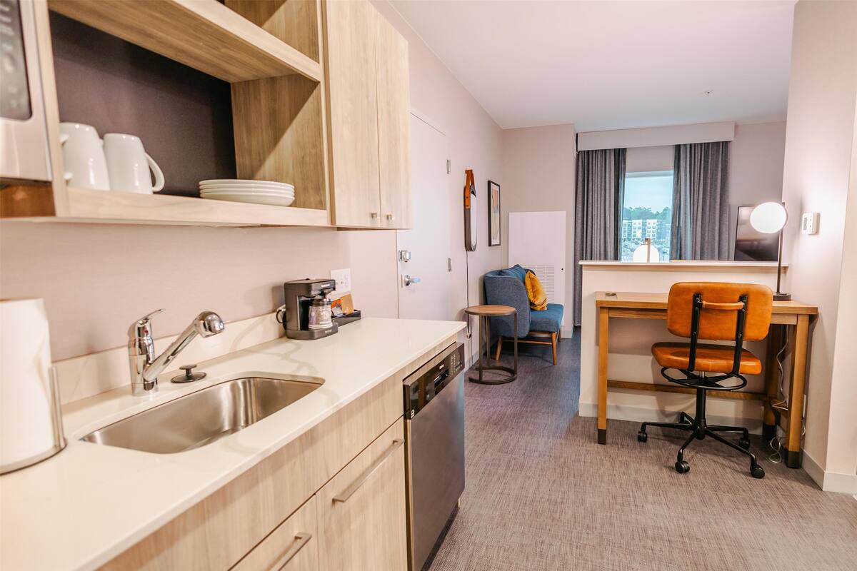 A compact hotel room with a kitchenette, a desk with a chair, a small round table, and a view of the city through the window.
