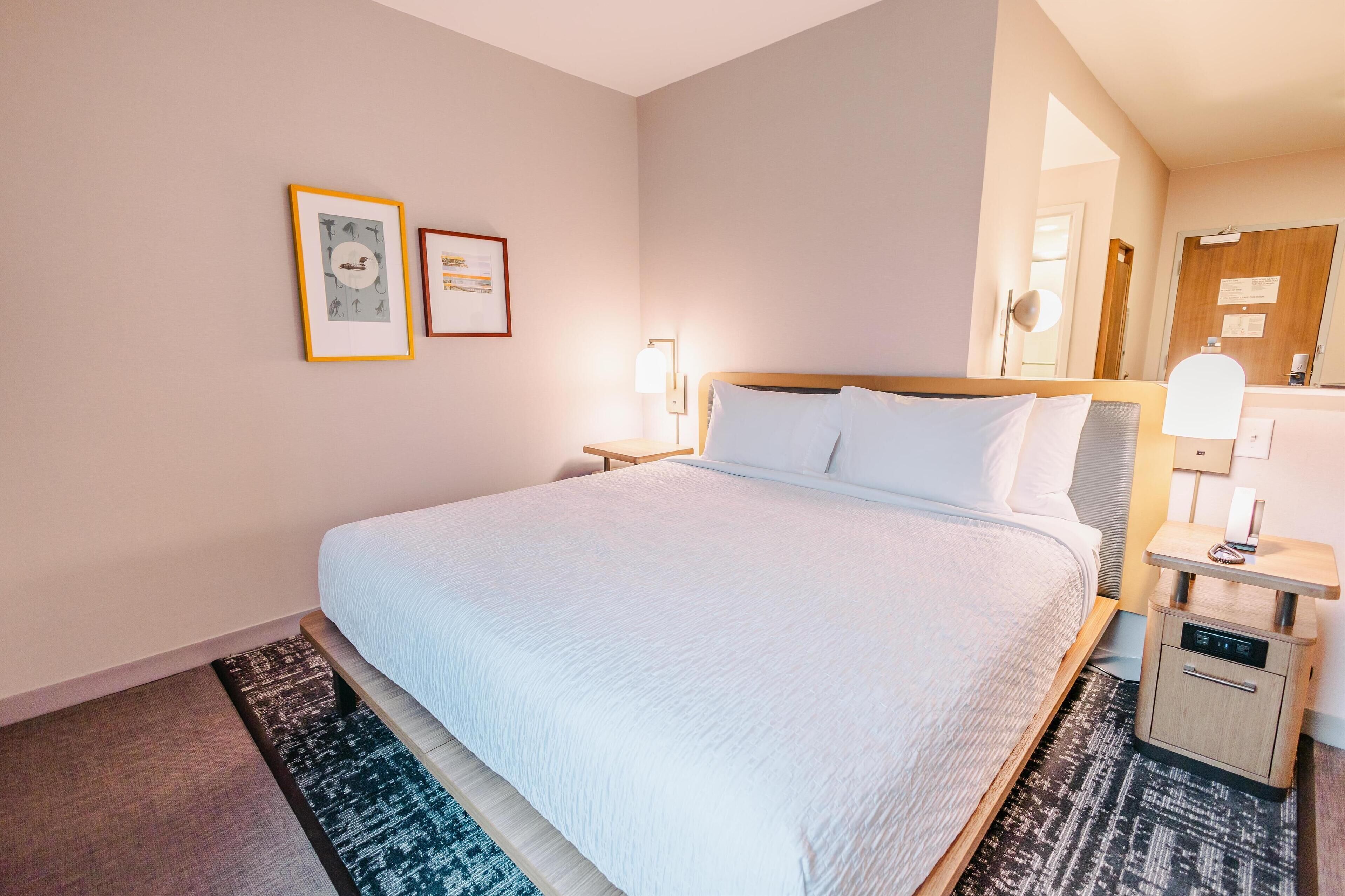 Quarto de hotel com uma cama grande, uma mesa de cabeceira com abajur e duas quadros na parede.