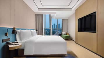 A modern hotel room with a large bed, a flat-screen TV, a desk with a phone, and a view of the cityscape through large windows.