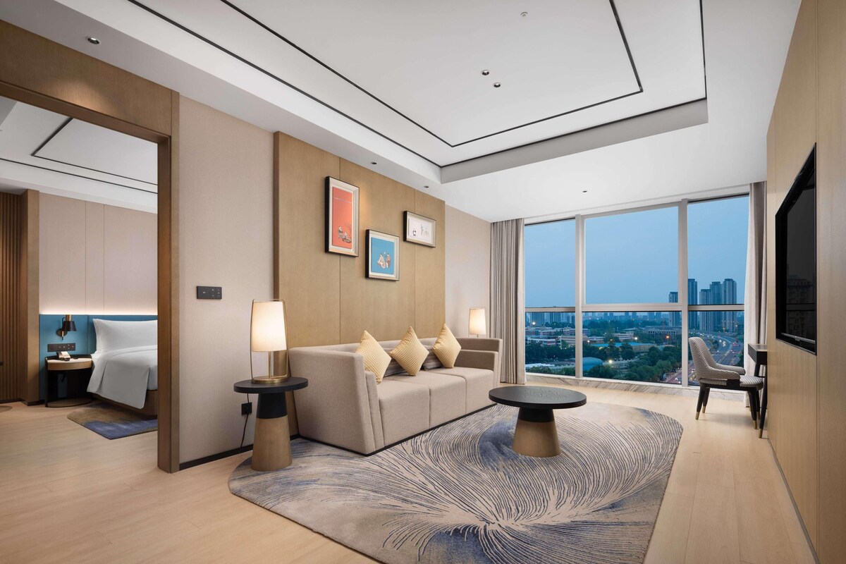 A modern hotel room with a large window, a sofa, a coffee table, and a TV.
