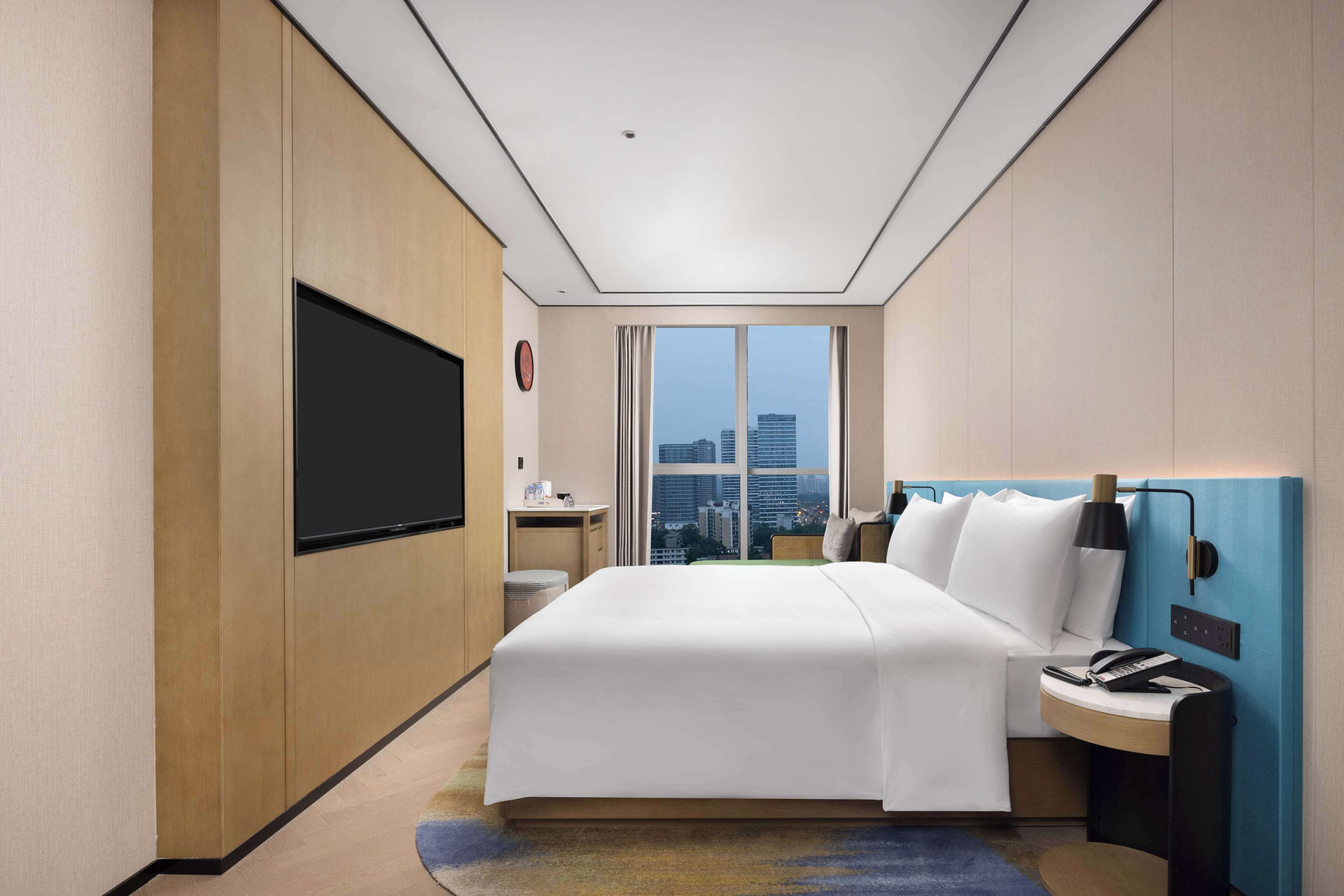 Una moderna camera d'hotel con un grande letto, una TV a schermo piatto montata a parete, una scrivania con sedia e vista sulla città attraverso ampie finestre.