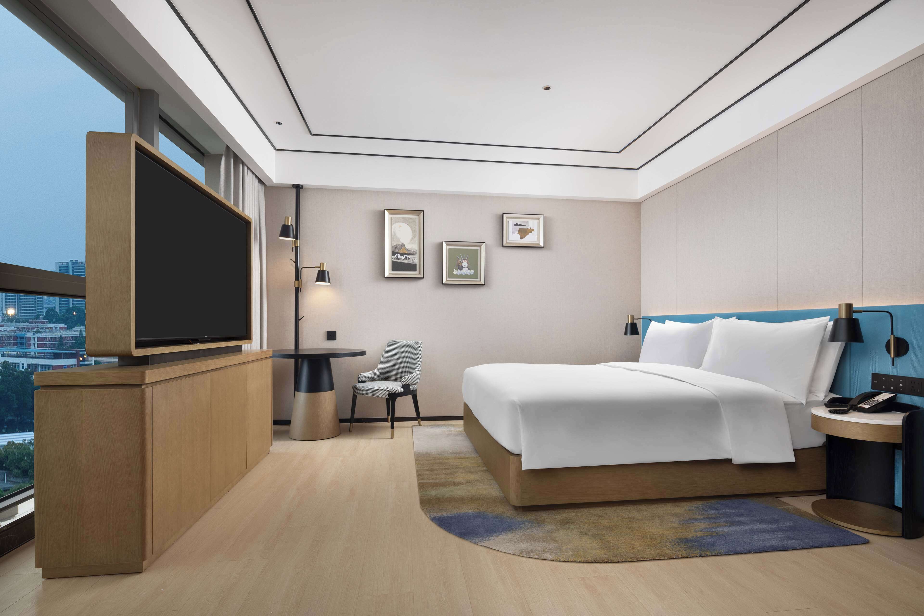 A modern hotel room with a large bed, a TV on a wooden console, a chair, and framed pictures on the wall.