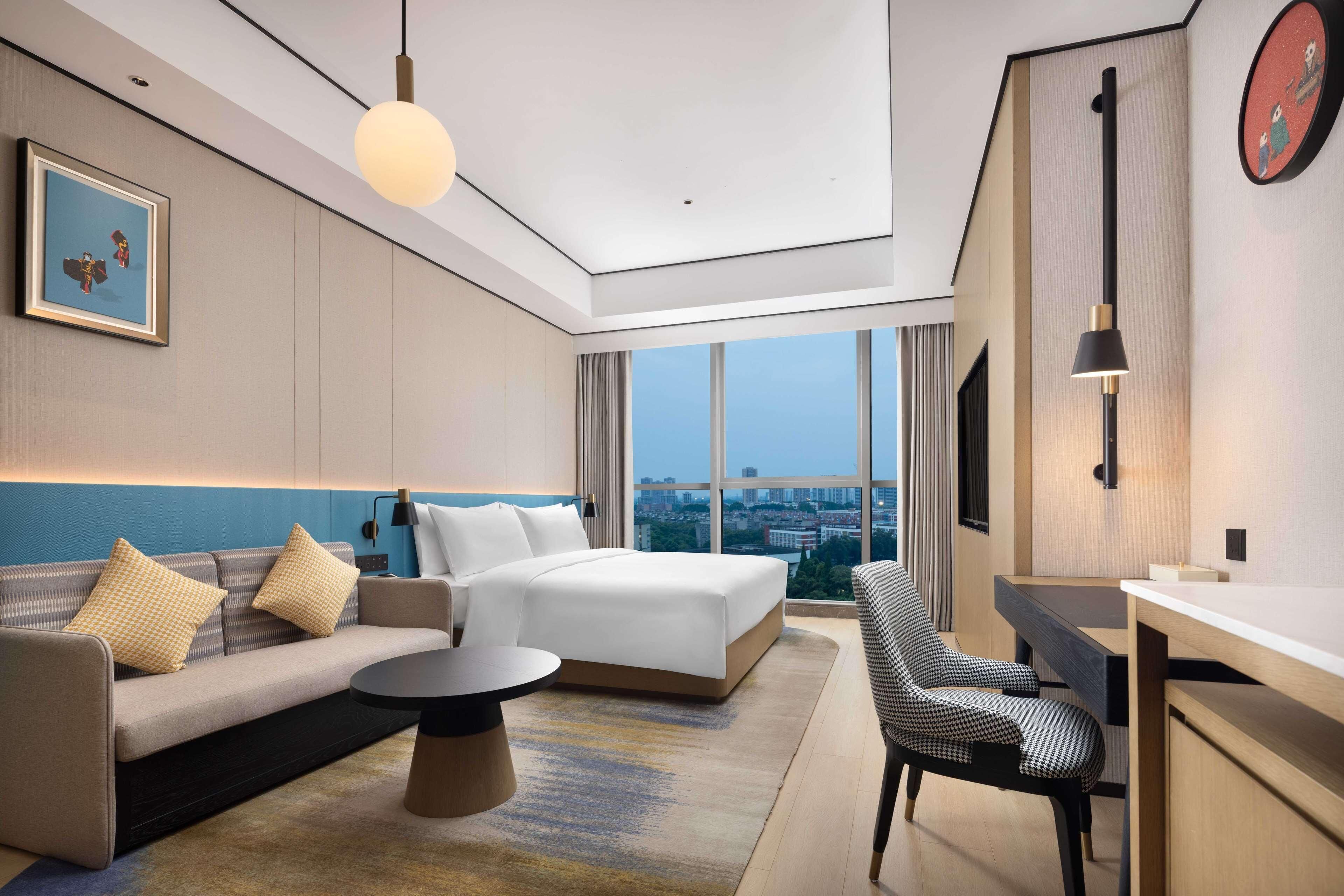 A modern hotel room with a large bed, a sofa, a dining table, and a view of the city.