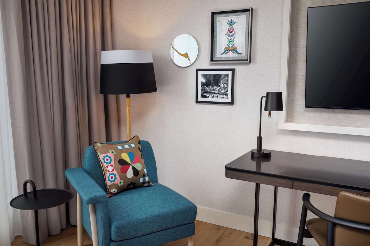 Een moderne kamer met een blauwgroene fauteuil, een klein rond tafeltje, een bureau en een grote flatscreen televisie aan de muur.