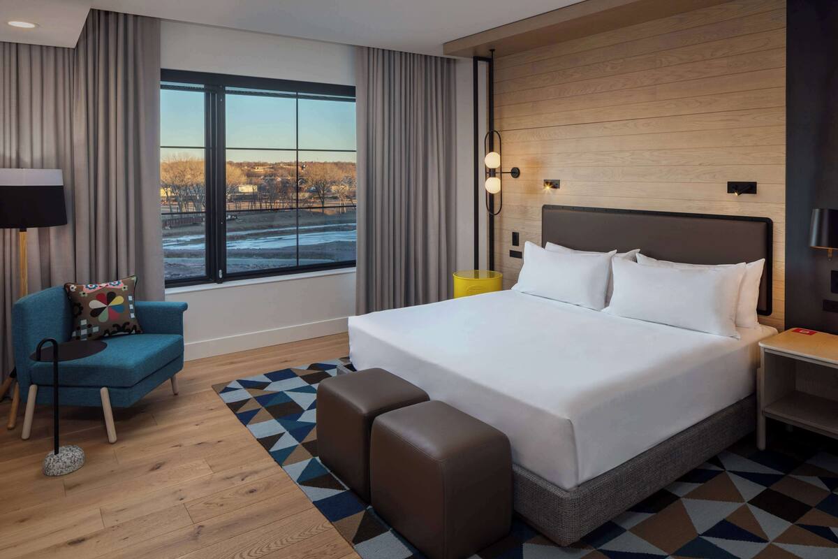 Een moderne hotelkamer met een groot bed, een blauwe fauteuil, een klein tafeltje en uitzicht op een besneeerd landschap.