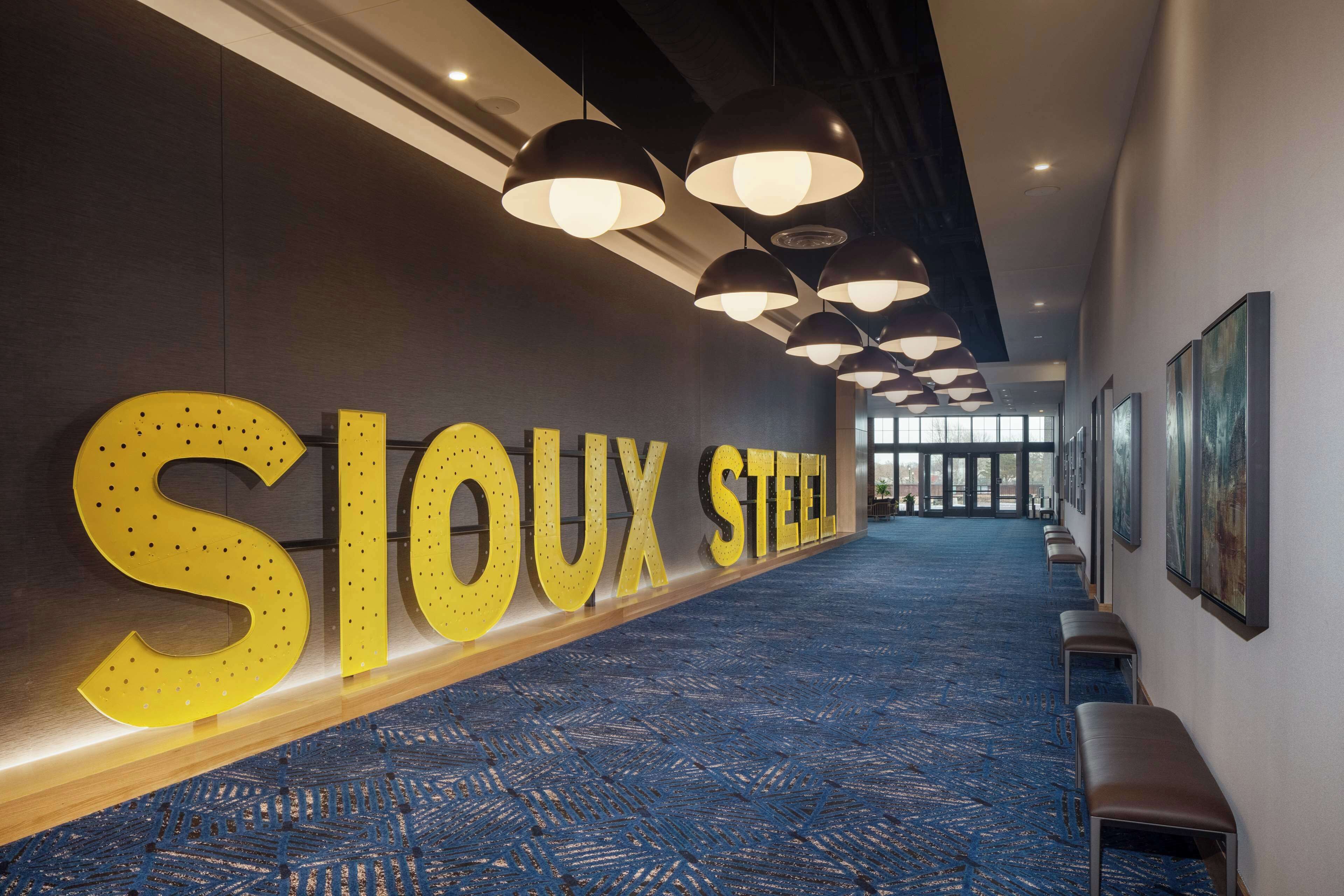 A hotel corridor with a blue carpet, large 'SIOUX STEEL' signage, and multiple pendant lights.