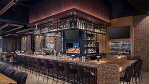 A bar area with a long counter, multiple stools, and shelves stocked with bottles. - Canopy By Hilton Sioux Falls Downtown (Sioux Falls)