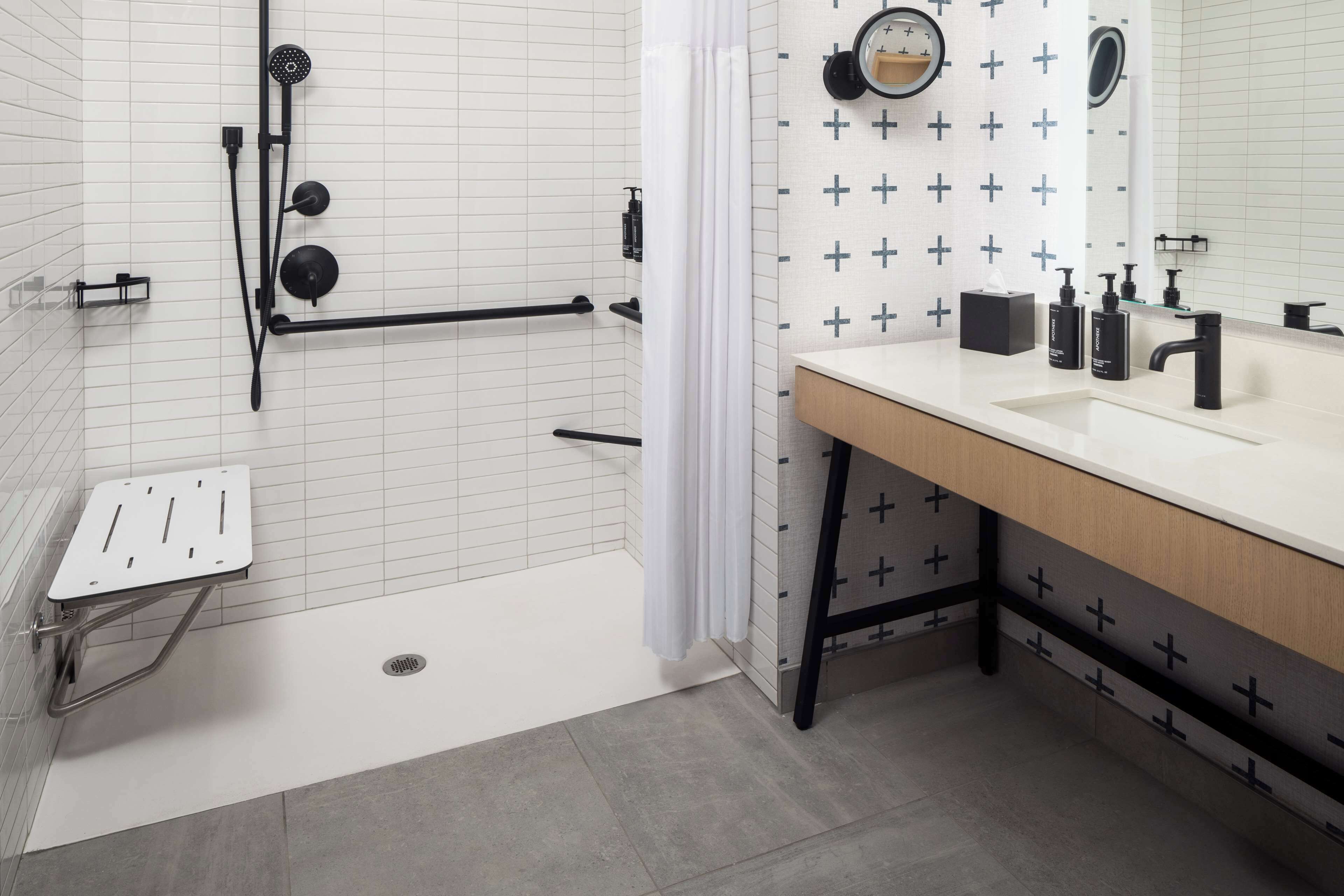 A modern bathroom with a white tiled shower area, a white sink with black fixtures, and a mirror with a clock.