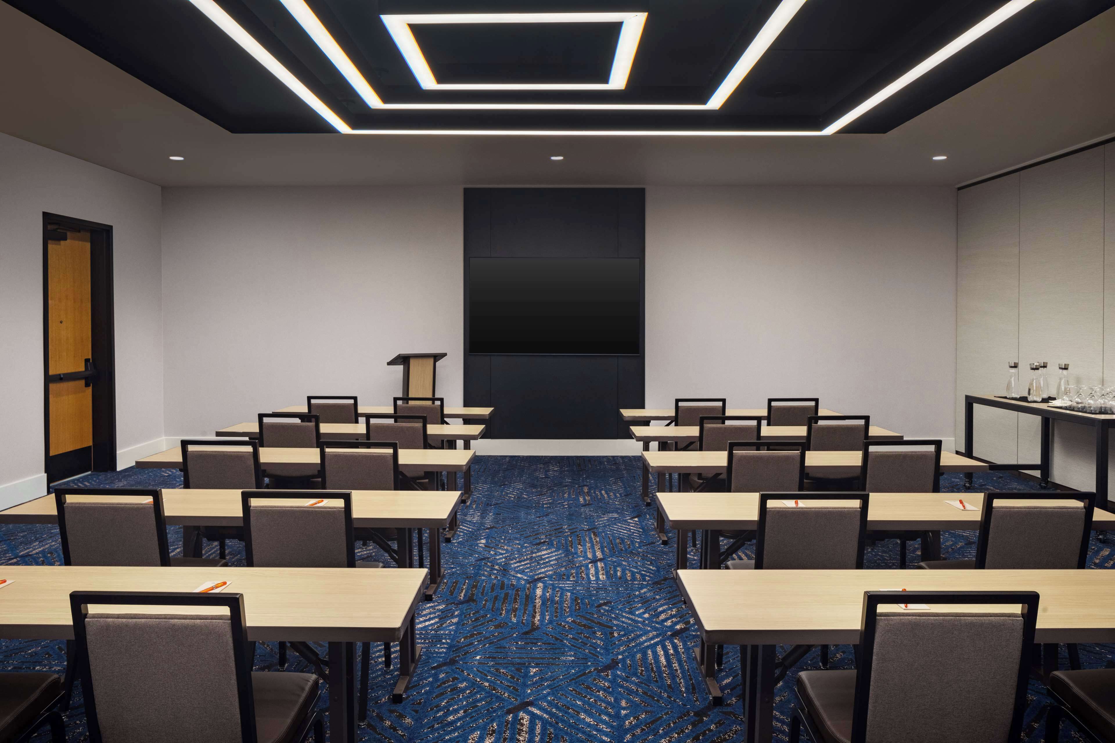 Een vergaderzaal met een lange tafel, stoelen, een spreekgestalte en een groot scherm.