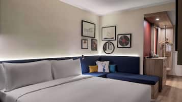A modern hotel room with a large bed, a sofa, and a desk. There are framed pictures on the wall and a small cabinet with a hanging rack.