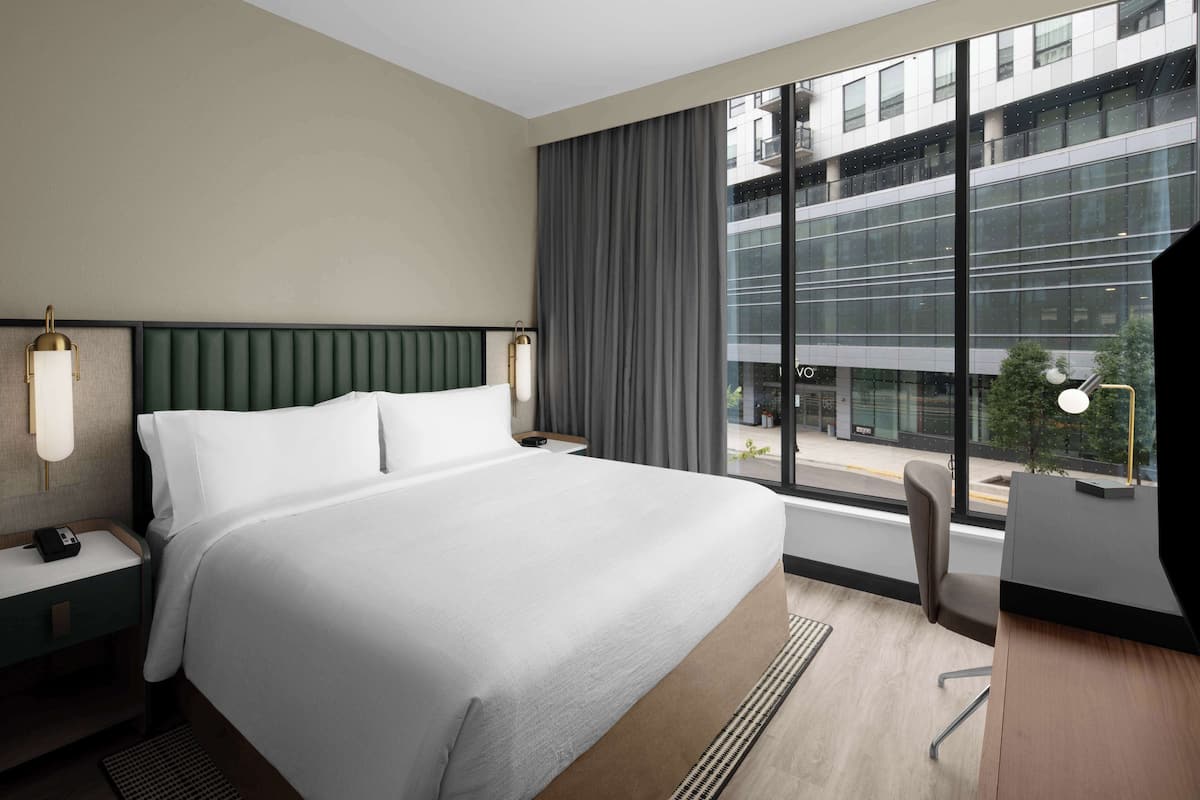 A modern hotel room with a large bed, a desk, and a view of a cityscape through large windows.