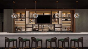 A bar area with a long counter, multiple shelves with bottles, glasses, and a flat-screen TV. There are four bar stools with green cushions. - Embassy Suites By Hilton Madison Downtown (Madison)