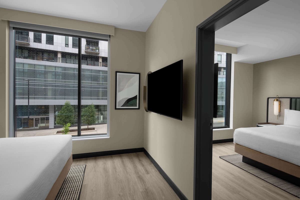 A modern hotel room with a large window, a flat-screen TV, a bed, and a bedside table.