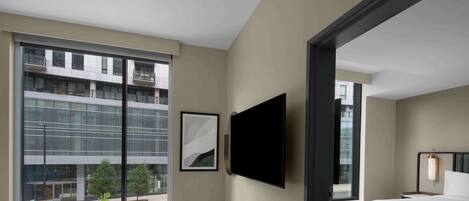 A modern hotel room with a large window, a flat-screen TV, a bed, and a bedside table.