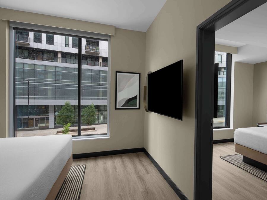 A modern hotel room with a large window, a flat-screen TV, a bed, and a bedside table.