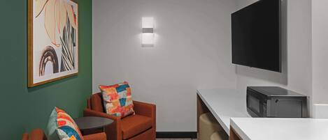 A compact room with a sofa, a small table, a microwave, and a flat-screen TV mounted on the wall.