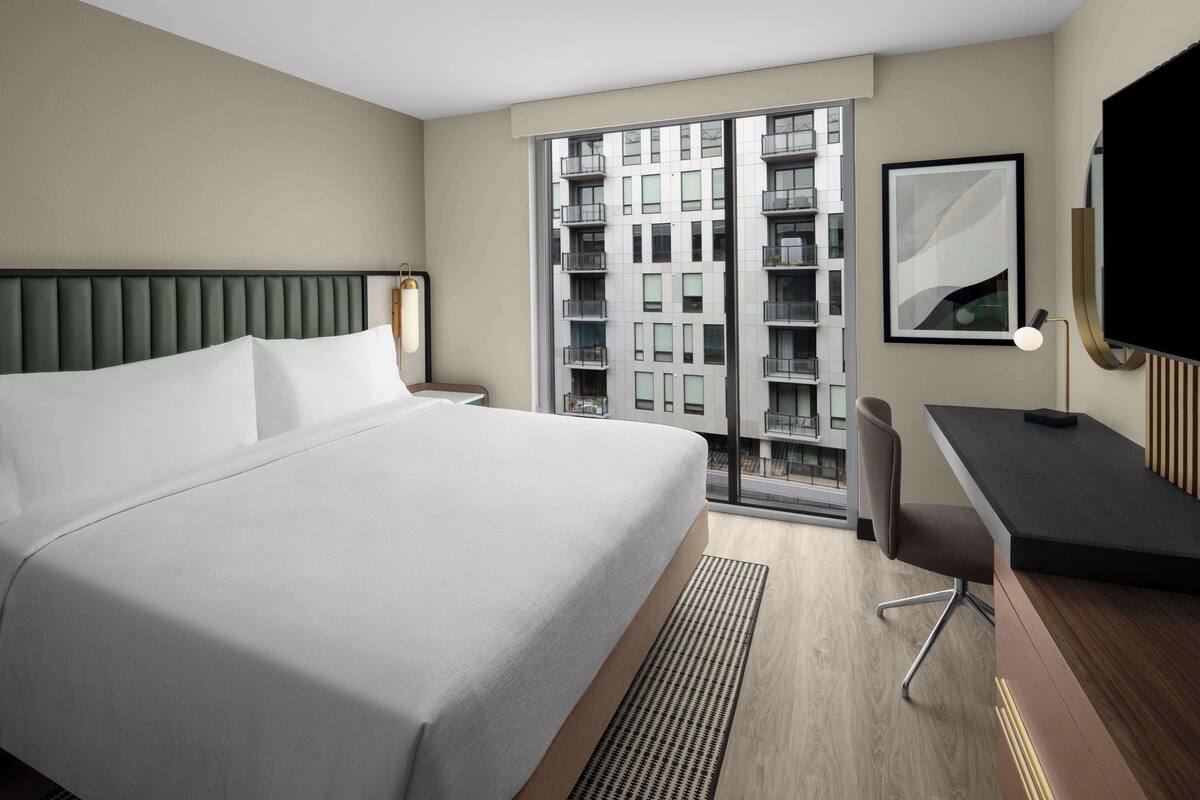 A modern hotel room with a large bed, a desk, a chair, a TV, and a window with a view of a building.