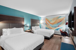 Habitación de hotel con dos camas, un escritorio, una silla, un televisor y un cuadro abstracto de colores en la pared.