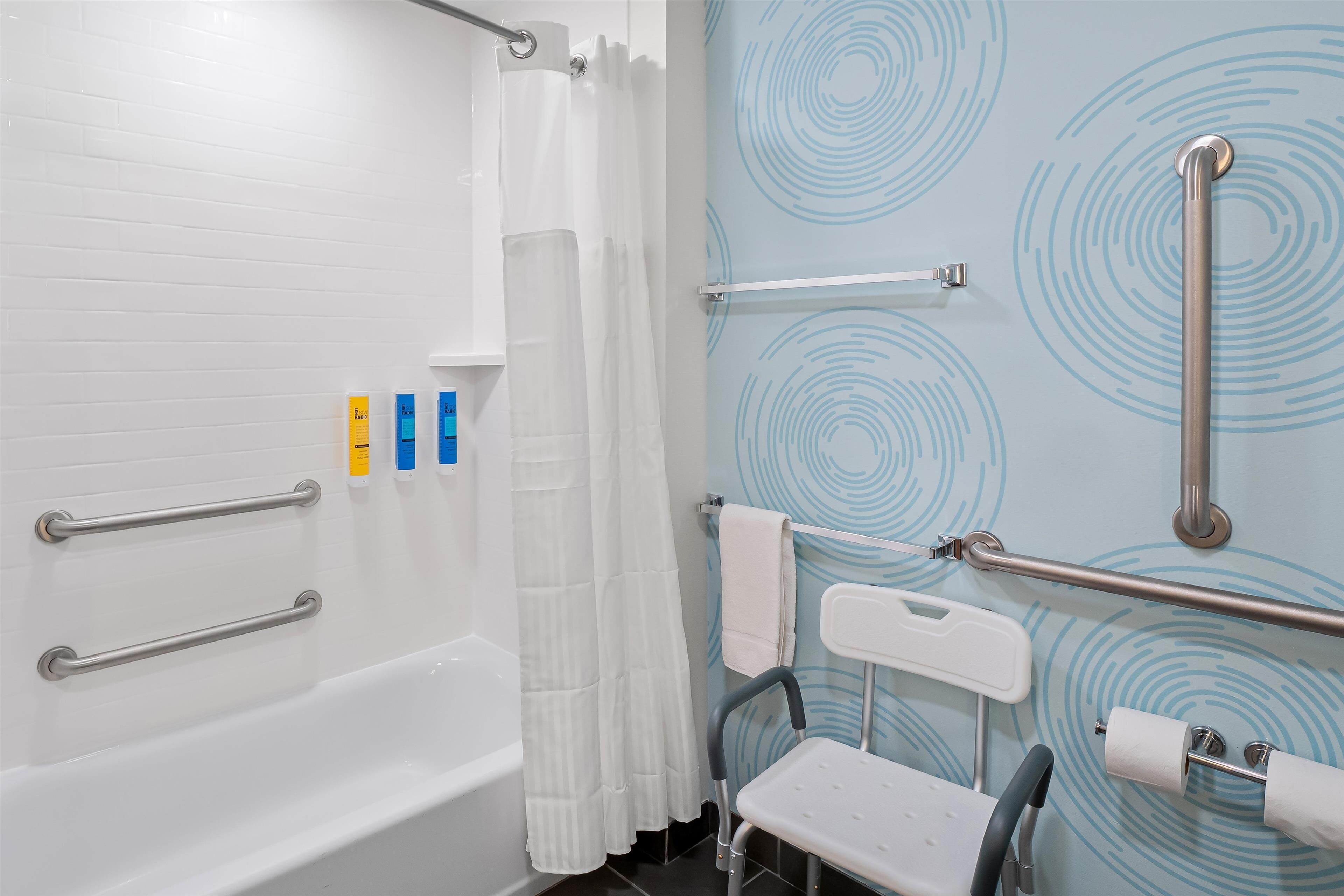 Un baño con una bañera blanca, una cortina de ducha, un pasamanos, una silla accesible para sillas de ruedas y un perchero para toallas.