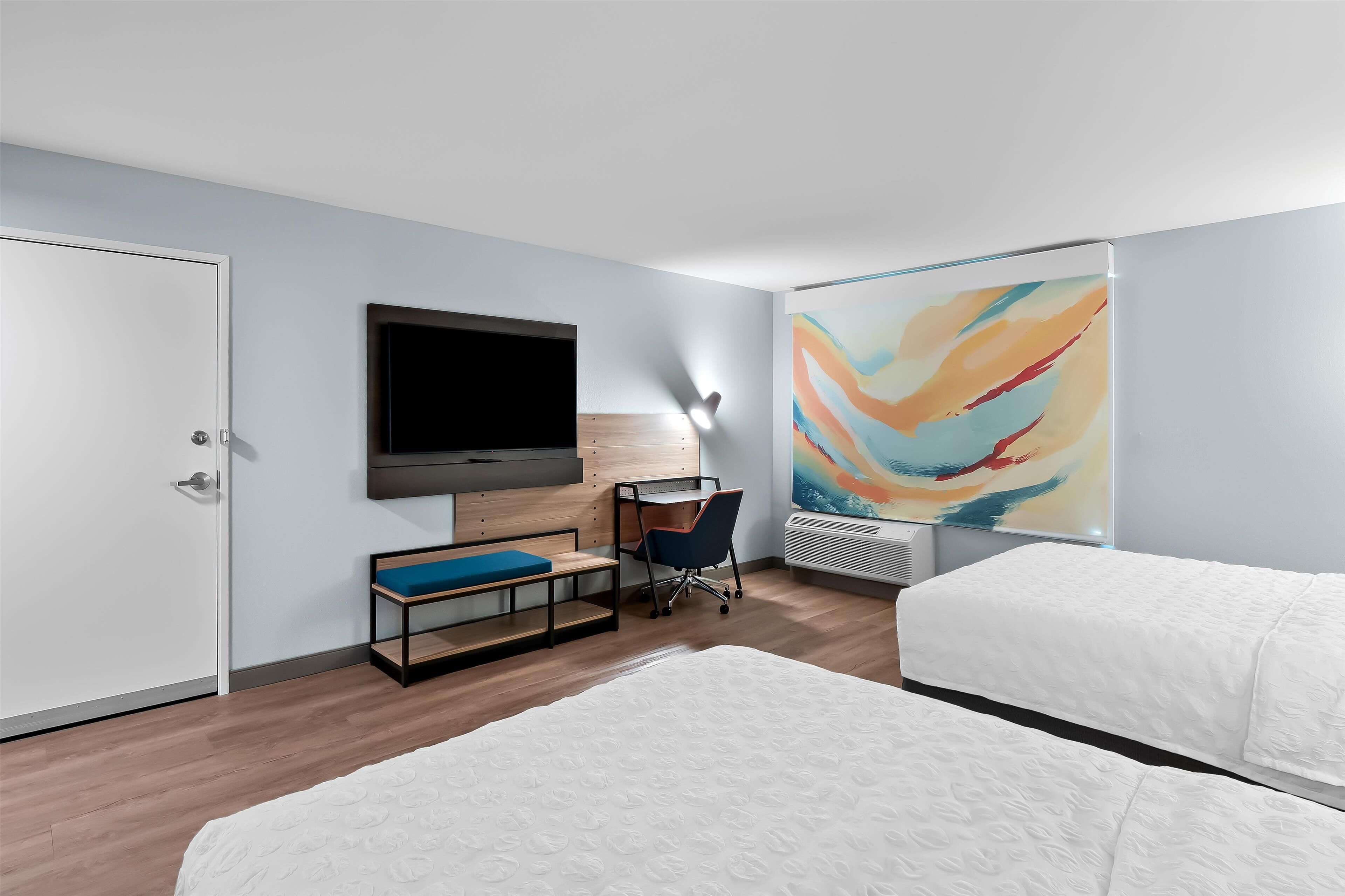 A hotel room with a bed, a desk with a chair, a television, and a large abstract painting on the wall.