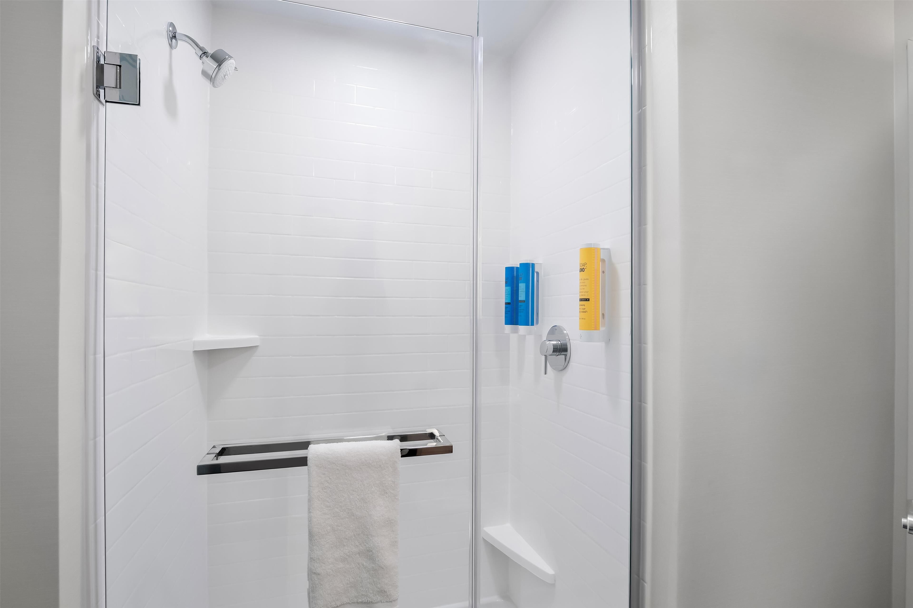 Un baño moderno con paredes de azulejos blancos, cabina de ducha de vidrio, un perchero y una repisa con artículos de aseo.