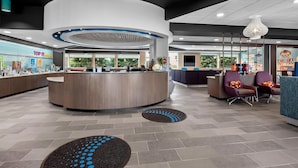 A modern reception area with a curved counter, seating with colorful cushions, and a large window providing a view of greenery outside. - Tru By Hilton Wauseon (Wauseon)
