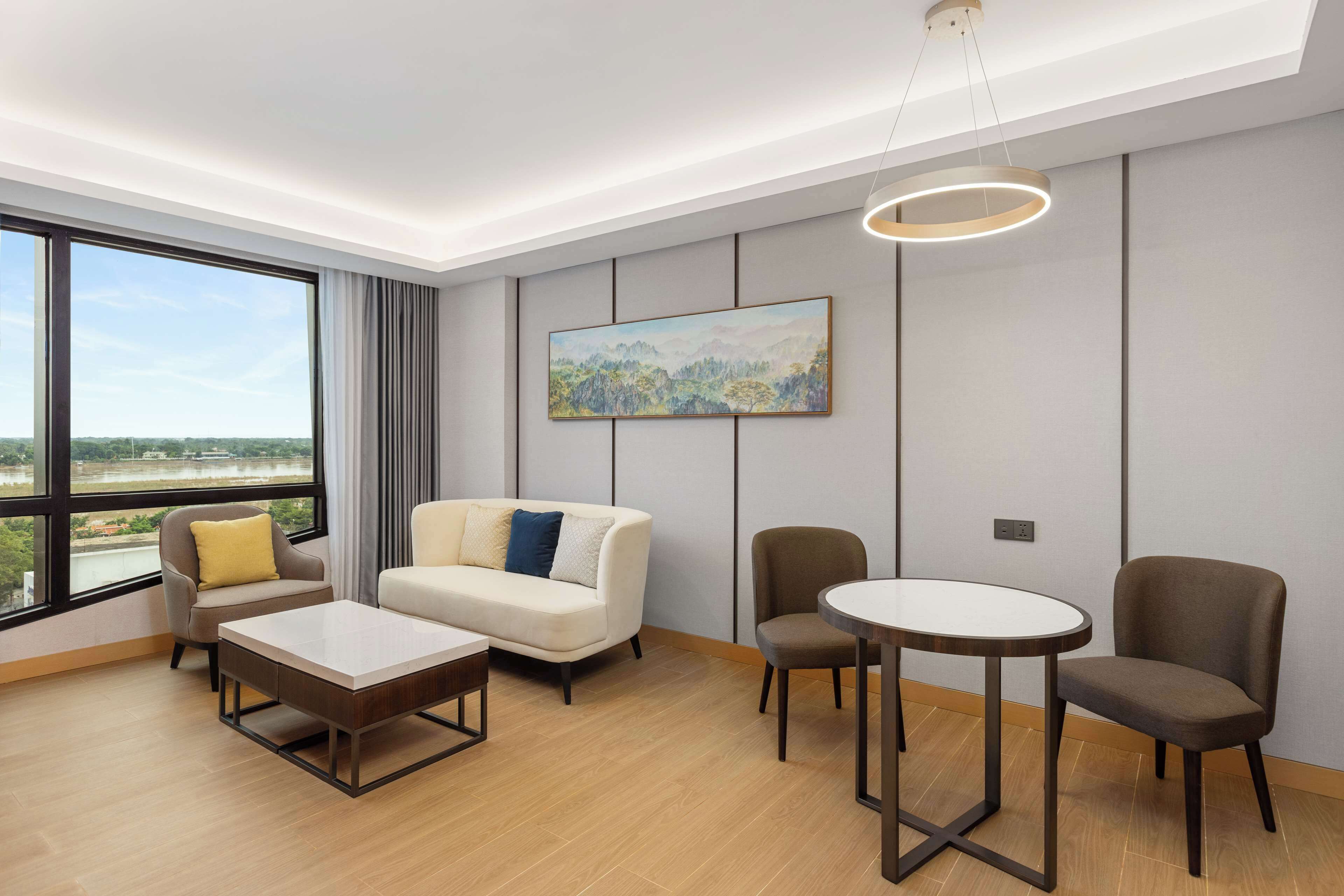 Une chambre d’hôtel moderne avec une grande fenêtre, un canapé, une table basse, une table ronde et un tableau au mur.