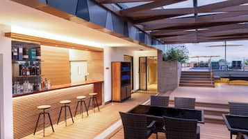 Un área de bar moderna al aire libre con suelo de madera, taburetes, mobiliario de mimbre y cerramiento de vidrio.