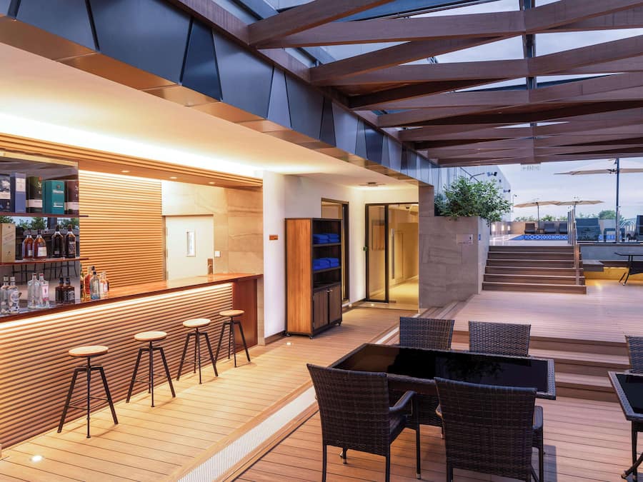 Un área de bar moderna al aire libre con suelo de madera, taburetes, mobiliario de mimbre y cerramiento de vidrio.