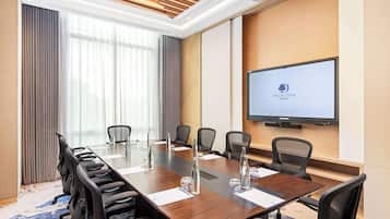 Een vergaderzaal met een lange tafel, stoelen, een projectorscherm en een groot raam met gordijnen.
