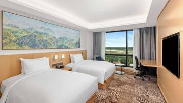A hotel room with two beds, a desk, a TV, and a large window offering a view of the outdoors.