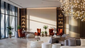 Un área de descanso moderna con un conjunto de sofás, dos sillones naranja y una mesa de centro. Hay plantas en macetas y un accesorio de iluminación decorativo.