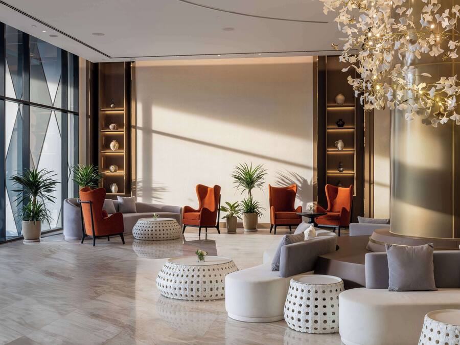 Un área de descanso moderna con un conjunto de sofás, dos sillones naranja y una mesa de centro. Hay plantas en macetas y un accesorio de iluminación decorativo.