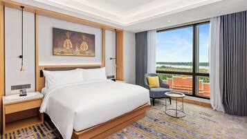 A hotel room with a large bed, a bedside table, a nightstand, a small round table, a chair, and a large window offering a view of the outdoors.