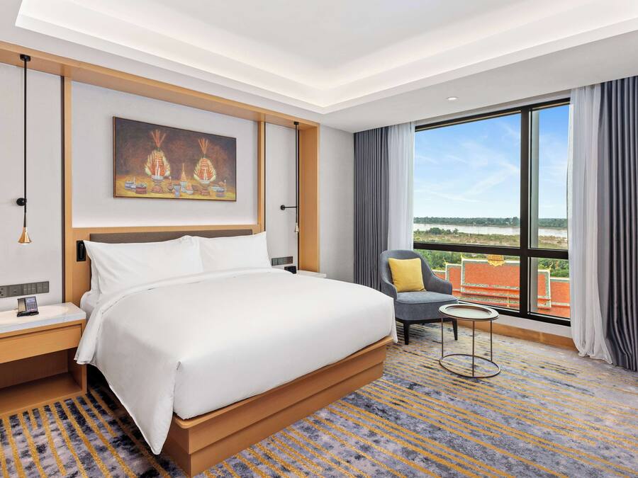 A hotel room with a large bed, a bedside table, a nightstand, a small round table, a chair, and a large window offering a view of the outdoors.