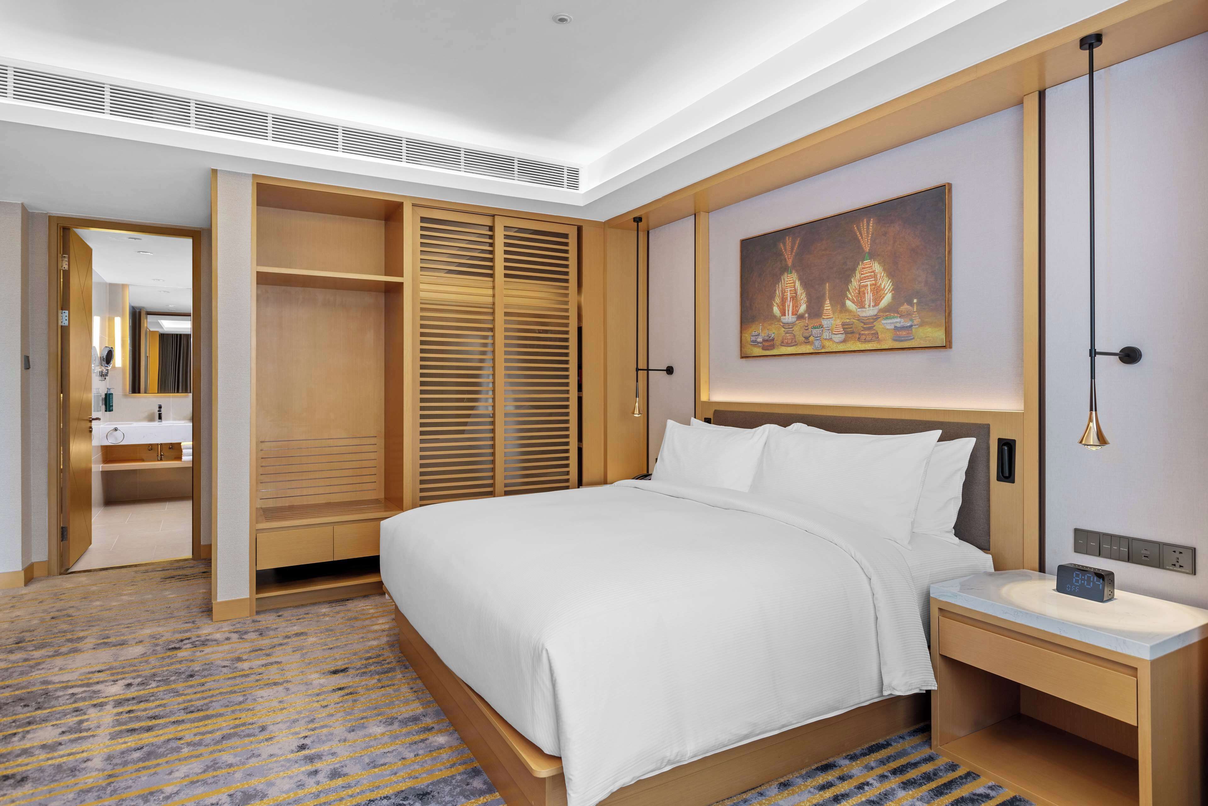 A hotel room with a large bed, wooden wardrobe, a bedside table with a lamp, and a painting on the wall.