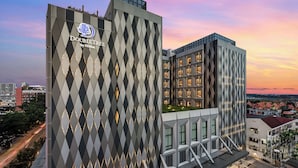 Un hotel moderno con fachada geométrica y ventanas iluminadas, rodeado de un paisaje urbano al anochecer.