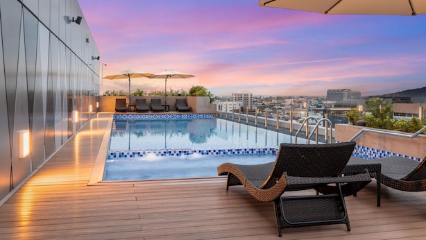 Zona de piscina en la azotea con sillones y sombrillas, con vistas a un paisaje urbano al atardecer.