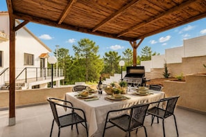 Patio esterno con tavolo apparecchiato per consumare pasti, una griglia barbecue e vista su un edificio residenziale.