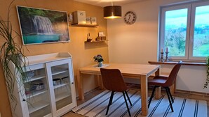 A dining area with a wooden table, two chairs, a kitchen cabinet, a clock, and a window with a view. - Ferienwohnung im Allgäu mit Direktem Blick auf den Hochgrat in Ruhiger Lage! (Argenbühl)