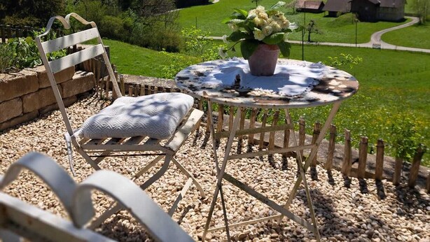 A round table with a potted plant, two white cushioned chairs, and a view of a green landscape with mountains in the distance. - Ferienwohnung im Allgäu mit Direktem Blick auf den Hochgrat in Ruhiger Lage! (Argenbühl)