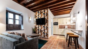 Una sala de estar con un sofá, una mesa de centro y un televisor montado en una pared con diseño geométrico. Hay una zona de cocina con microondas, un fregadero y taburetes.