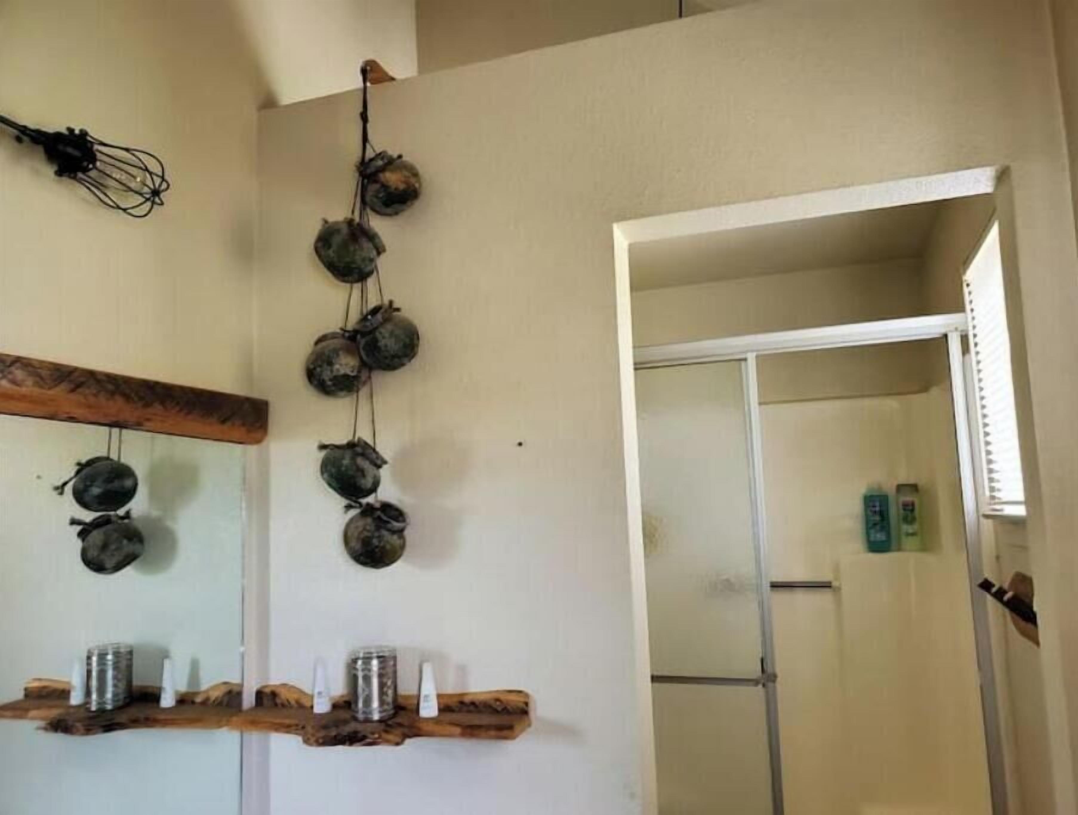 A bathroom with a wooden shelf holding toiletries, a hanging decoration of round objects, and a mirror above the shelf.