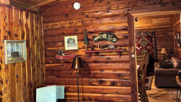 Interior de una cabaña de troncos con una zona de estar acogedora, una pequeña mesa y una escalera que lleva a otra habitación.
