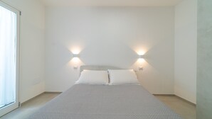A minimalist bedroom with a large bed, two wall-mounted lights, and a sliding glass door. - Relax Flatroom 22 (Olbia)