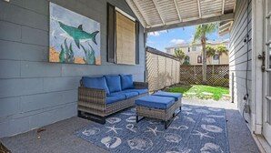 Une terrasse couverte avec un canapé et un pouf en osier, un tapis décoratif aux motifs marins et un tableau représentant un requin accroché au mur.