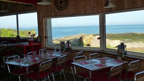 Ein Restaurant mit Holzwänden und großen Fenstern mit Blick auf das Meer. Die Tische sind mit roten Stühlen und weißen Tischdecken gedeckt.